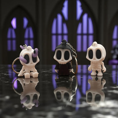 Drei 3D-gedruckte Schlüsselanhänger: Skelett-Blob (weiß/lila), Grim-Reaper in Robe und Skelett-Blob (weiß) mit Schlüsselring auf dunklem Tisch – Halloween Produktfoto.