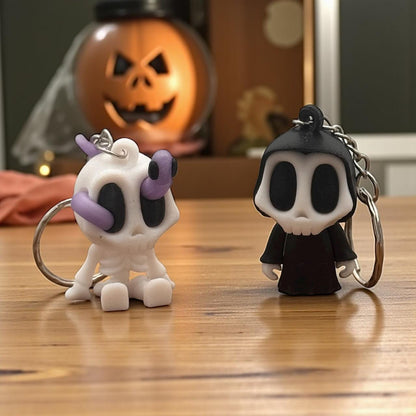 Set-Foto: Skelett-Blob (weiß/lila) und Grim-Reaper-Schlüsselanhänger zusammen auf Holztisch; im Hintergrund Kürbis-Deko (Jack-o’-Lantern), herbstliche Stimmung.